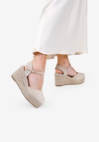 Sandalias de cuña beige con un upper de tela texturizada, plataforma de yute trenzado y correa para el tobillo con cierre de hebilla.