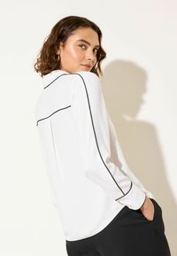 Biela košeľa s dlhým rukávom s čiernym lemovaním pozdĺž ramien a rukávov, s voľným strihom a detailom záhybu na chrbte.