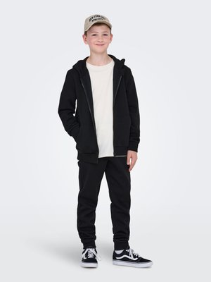 Felpa nera con zip sopra una maglietta bianca, abbinata a pantaloni della tuta neri e sneakers nere. Indossa anche un cappellino beige con la scritta "NEW YORK CITY".