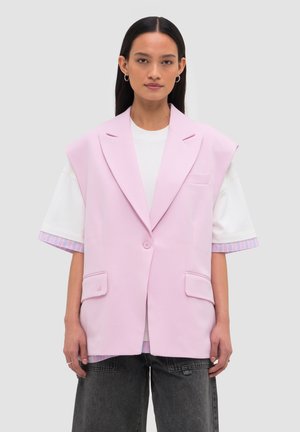 Frau mit glatten schwarzen Haaren trägt einen übergroßen pastellrosa ärmellosen Blazer über einem weißen Hemd und schwarzen Jeans, stehend vor schlichtem Hintergrund.