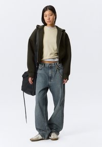 Νεαρή γυναίκα με φαρδύ φούτερ με φερμουάρ σε ελιά πράσινο, μπεζ κροπ τοπ, φαρδιά τζιν και λευκά αθλητικά παπούτσια, κρατώντας μια μαύρη τσάντα πλάτης στο πλάι της.