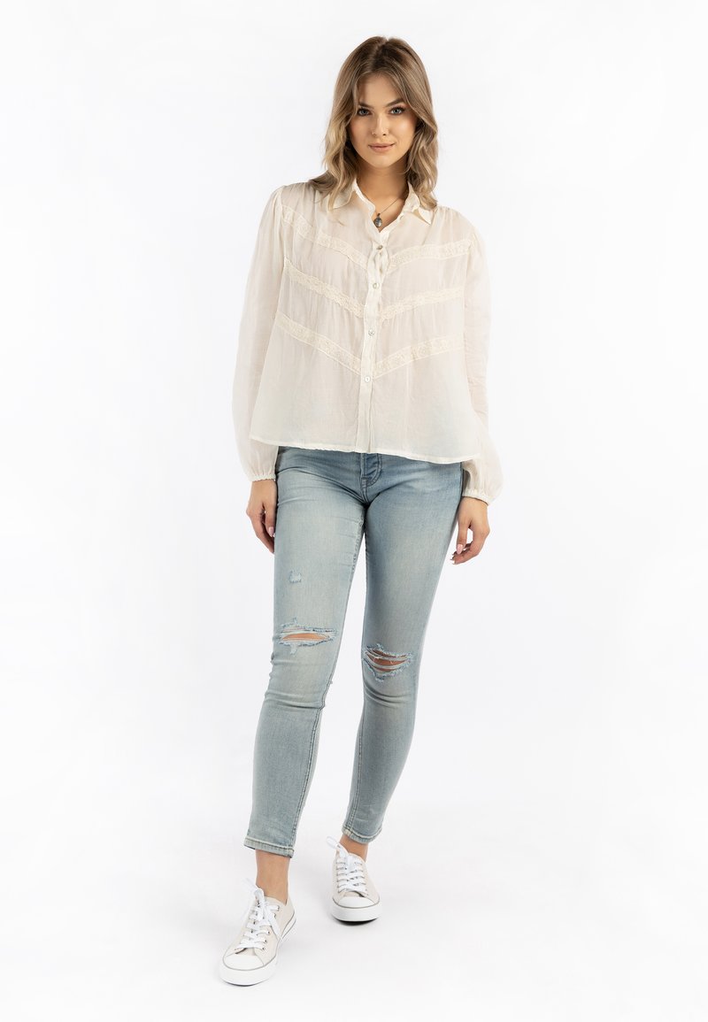 Blusa bianca e trasparente con dettagli in pizzo, abbinata a jeans strappati azzurri chiari e sneakers bianche. La blusa presenta una chiusura a bottoni frontale e maniche a palloncino.