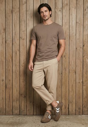 Mann steht vor einer Holzwand, trägt ein braunes T-Shirt, beige Hose und braune Sneaker mit weißen Chevron-Streifen, eine Hand in der Tasche.