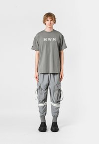 Camiseta gris de manga corta con el logo "MWM", combinada con pantalones cargo grises con cremalleras y acentos blancos, y calzado negro texturizado.