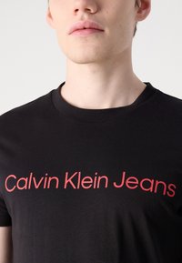 Osoba koja nosi crnu majicu s okruglim izrezom s natpisom "Calvin Klein Jeans" ispisanim velikim crvenim slovima preko prsa.