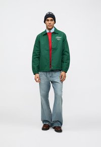 Πράσινο αδιάβροχο μπουφάν με κλείσιμο από κουμπιά, στρωμένο πάνω από ένα κόκκινο πουλόβερ, ανοιχτό μπλε πουκάμισο και ανοιχτό μπλε φαρδύ τζιν. Μαύρο σκουφάκι με σχέδιο.