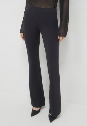 Pantaloni neri a zampa d'elefante con una texture liscia e cintura elastica, abbinati a décolleté nere a punta. Leggero riflesso morbido dalla luce circostante.