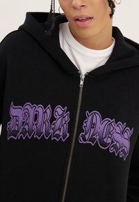 Personne portant un sweat à capuche noir zippé avec le texte gothique violet "DARKNESS" sur un t-shirt blanc, visage partiellement visible avec une boucle d'oreille anneau.