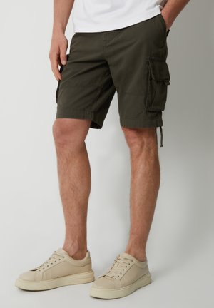Olivegrüne Cargoshorts für Herren mit Seitentaschen, getragen mit beigen Low-Top-Sneakern, stehend vor einem schlichten hellen Hintergrund.