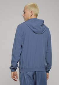 Kék sportdzseki kapucnival, rugalmas mandzsettákkal és derékpánttal, könnyű anyagból készült, sima textúrával.