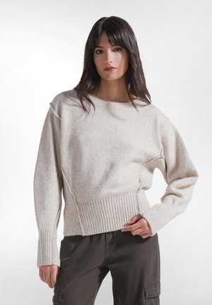 Kvinde med mørkt hår iført en lysebeige strikket sweater med ribbet kant og manchetter, kombineret med mørke bukser, stående mod en ensfarvet baggrund.