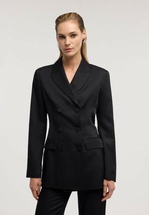 Mujer que lleva un blazer negro entallado de doble botonadura con solapas con muesca y bolsillos con solapa en la parte frontal sobre un fondo claro y liso.