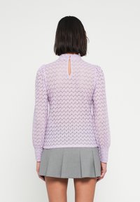 Πλεκτό τοπ σε λιλά απόχρωση με υφασμένο εξάγωνο μοτίβο, μακριά μανίκια και ένα άνοιγμα στο πίσω μέρος. Συνδυάζεται με γκρι πλισέ φούστα.