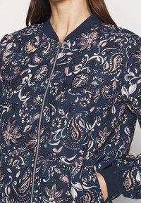 Marinblå bomberjacka med detaljerade blommiga och paisleymönster i rosa, vitt och grönt. Har dragkedjestängning och ribbad halsringning och ärmslut.