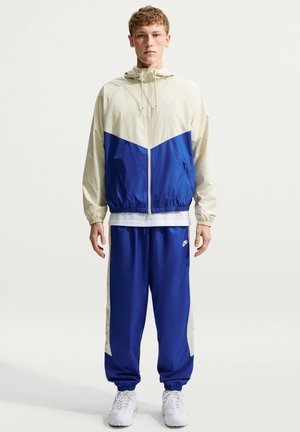 Ung mand står iført en blå og beige hættetrøje med lynlås, matchende blå træningsbukser med beige striber og hvide sneakers.