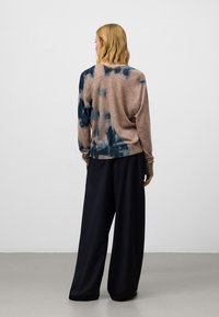 Pull en maille marron avec motifs tie-dye bleus, encolure ronde et manches longues, associé à un pantalon large noir et ample.