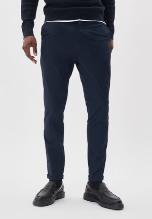 Pantaloni blu navy con una texture liscia, slim fit e orli arrotolati. Abbinati a mocassini neri, caratterizzati da una superficie texturizzata e suole spesse.