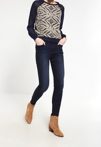 Marinblå och krämfärgad topp med blommönster och långa genomskinliga ärmar, matchad med mörka skinny jeans och ljusa ankelstövlar med klack.