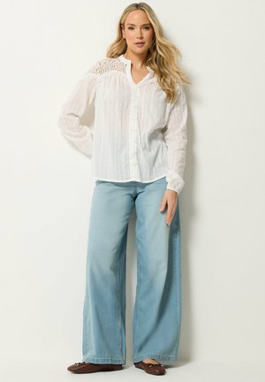 Giovane donna con lunghi capelli biondi che indossa una camicetta bianca con spalle in pizzo, jeans a gamba larga color azzurro chiaro e scarpe basse marroni, in piedi contro un muro senza decorazioni.