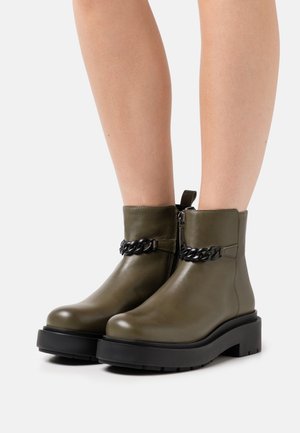 Stivaletti in pelle verde oliva al collo del piede con suole nere spesse e dettagli in catena nera, indossati su gambe nude su uno sfondo bianco.