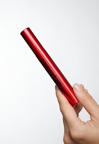 Tube de mascara en aluminium rouge avec un design effilé, portant un texte noir disant "SuperFan". Une main tenant le tube devant un fond clair.