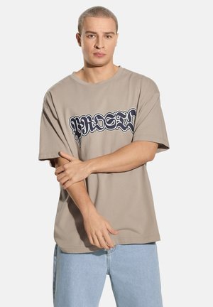 Beżowa bawełniana koszulka o luźnym kroju, z ciemnoniebieskim nadrukiem graficznym na wysoko ści klatki piersiowej. Noszona z jasnoniebieskimi dżinsami.