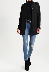Mujer que lleva un abrigo negro de manga larga abotonado, top negro, vaqueros azules desgastados con dobladillos deshilachados, botas negras hasta el tobillo y un cinturón decorativo.