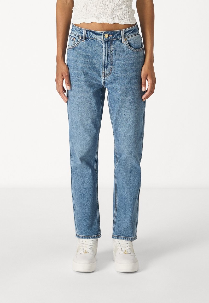 Femme portant un jean bleu taille haute à jambe droite, des baskets blanches et un top blanc texturé court sur un fond uni.