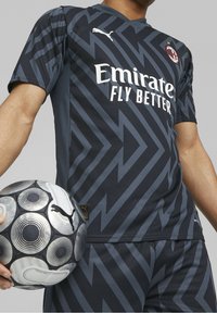 Tmavě šedý fotbalový dres s geometrickým zigzagovým vzorem, který má bílé logo a akcenty. Hráč drží fotbal s černo-stříbrným vzorem.