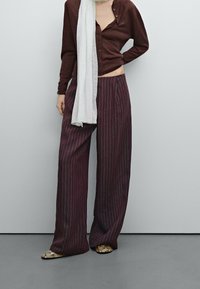 Bordeaux wijde pantalon met verticale witte strepen, gecombineerd met een donker vest met knopen en een lichte sjaal over de schouders gedrapeerd.