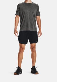 Man som bär en mörkgrå t-shirt med kort ärm, svarta träningsshorts och svarta löparskor står mot en vit bakgrund.