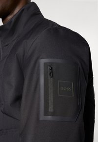 Μαύρο υφασμάτινο μπουφάν με φερμουάρ σε τσέπη στον πάνω βραχίονα και λάστιχο ετικέτα που γράφει "BOSS." Το υλικό φαίνεται ανθεκτικό και λειτουργικό.