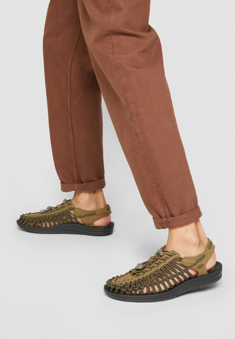 Brune sandaler med vevet overdel, gummisåle og justerbare stropper. Kombinert med brune oppbrettede bukser. Uformelt fottøy for utendørs bruk.