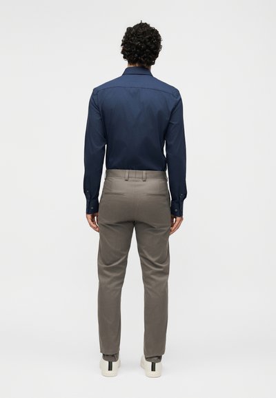 Άντρας με σγουρά μαλλιά, που φοράει μπλε πουκάμισο με μακριά μανίκια, παντελόνι ταουπ και λευκά αθλητικά παπούτσια, στέκεται γυρισμένος πλάτη σε απλό λευκό φόντο.