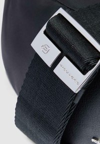 Bolsa negra con un exterior sintético liso, correa texturizada y herrajes metálicos con detalle de marca. Diseñada funcionalmente para la durabilidad.