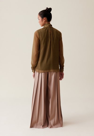 Blusa de manga larga verde oliva con espalda plisada y cuello alto, combinada con pantalones anchos beige con pliegues verticales.