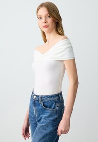 Weißer, gerippter, schulterfreier Bodysuit mit V-Ausschnitt, kombiniert mit hochgeschnittenen blauen Jeans aus Denim im klassischen Schnitt und mit Taschen.