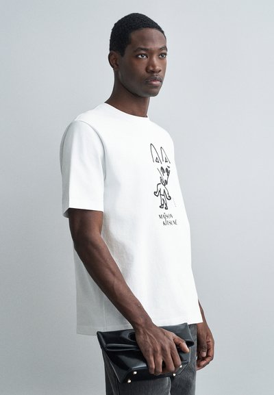 Mann trägt ein weißes T-Shirt von Maison Kitsuné und hält eine schwarze Leder-Clutch-Tasche vor einem schlichten grauen Hintergrund.