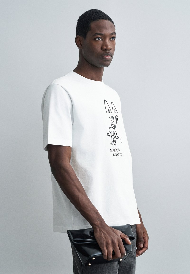 Muž v bílém tričku Maison Kitsuné drží černou koženou psaníčko před jednoduchým šedým pozadím.