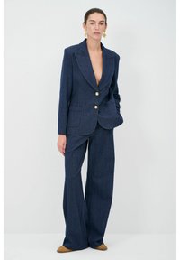 Completo in denim blu navy con giacca su misura dotata di bottoni dorati, rever a punta e tasca, abbinato a pantaloni a gamba larga.