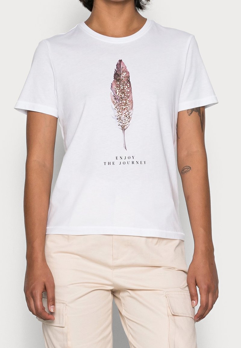 Personne portant un t-shirt blanc avec un graphisme de plume rose et le texte "Enjoy the Journey,", associé à un pantalon cargo beige clair.