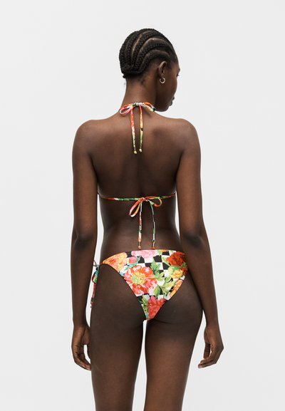 Γυναίκα με πλεξούδες που φοράει μπικίνι με λουλουδάτο μοτίβο και δεσίματα στον λαιμό, την πλάτη και τα ισχία, στέκεται μπροστά σε ένα απλό φωτεινό φόντο.