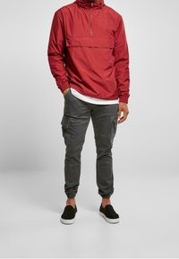 Mężczyzna w czerwonym kapturze, białej koszulce, ciemnoszarych dresowych spodniach cargo i czarnych butach wsuwanych stoi na tle jednolitym.