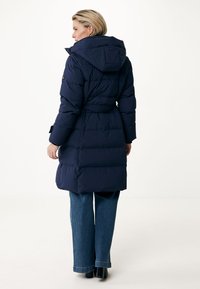 Cappotto puffo blu navy con vita cinchata, cappuccio e maniche lunghe. Il tessuto è liscio con una trama trapuntata. Abbinato a pantaloni di jeans a gamba larga.