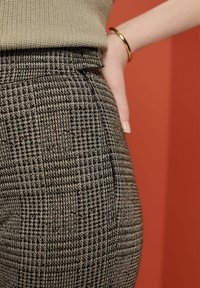 Gros plan sur une personne portant un pantalon taille haute à motif pied-de-poule et un pull en tricot beige, avec un bracelet doré visible au poignet.