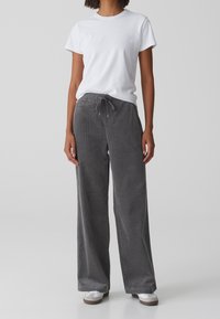Personne portant un t-shirt blanc uni à manches courtes, un pantalon large en velours côtelé gris avec une taille à cordon, et des chaussures blanches décontractées.