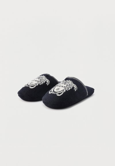 Μαύρες παντόφλες τύπου slip-on με λευκά κεντημένα σχέδια προσώπου Μέδουσας στην κορυφή, τοποθετημένες σε απλό φωτεινό φόντο.