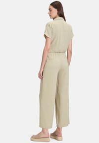 Beige jumpsuit med korte ærmer, krave, elastisk talje og vide ben. Fremstillet af et blødt, struktureret stof. Et par lyse sandaler er synlige.