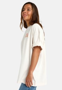 Svetlo krémové oversized tričko vyrobené z mäkkého bavlneného materiálu s krátkymi prehnutými rukávmi a malým viacfarebným grafickým dizajnom na prednej strane.