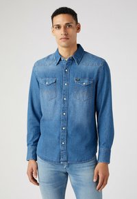 Camisa de mezclilla en azul medio con cierre de botones, cuello extendido, dos bolsillos delanteros y detalles de costura en contraste. Mangas largas, corte relajado.